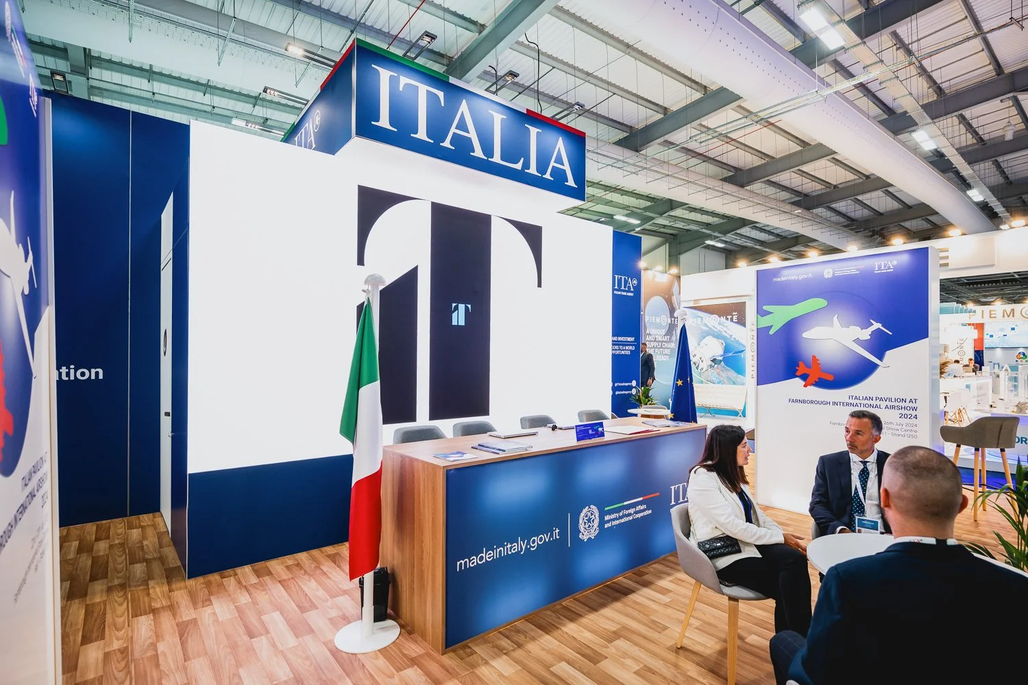 Booth at Farnborough International Airshow displaying Italy’s branding and flags, with two men and a woman conversing at a small table.