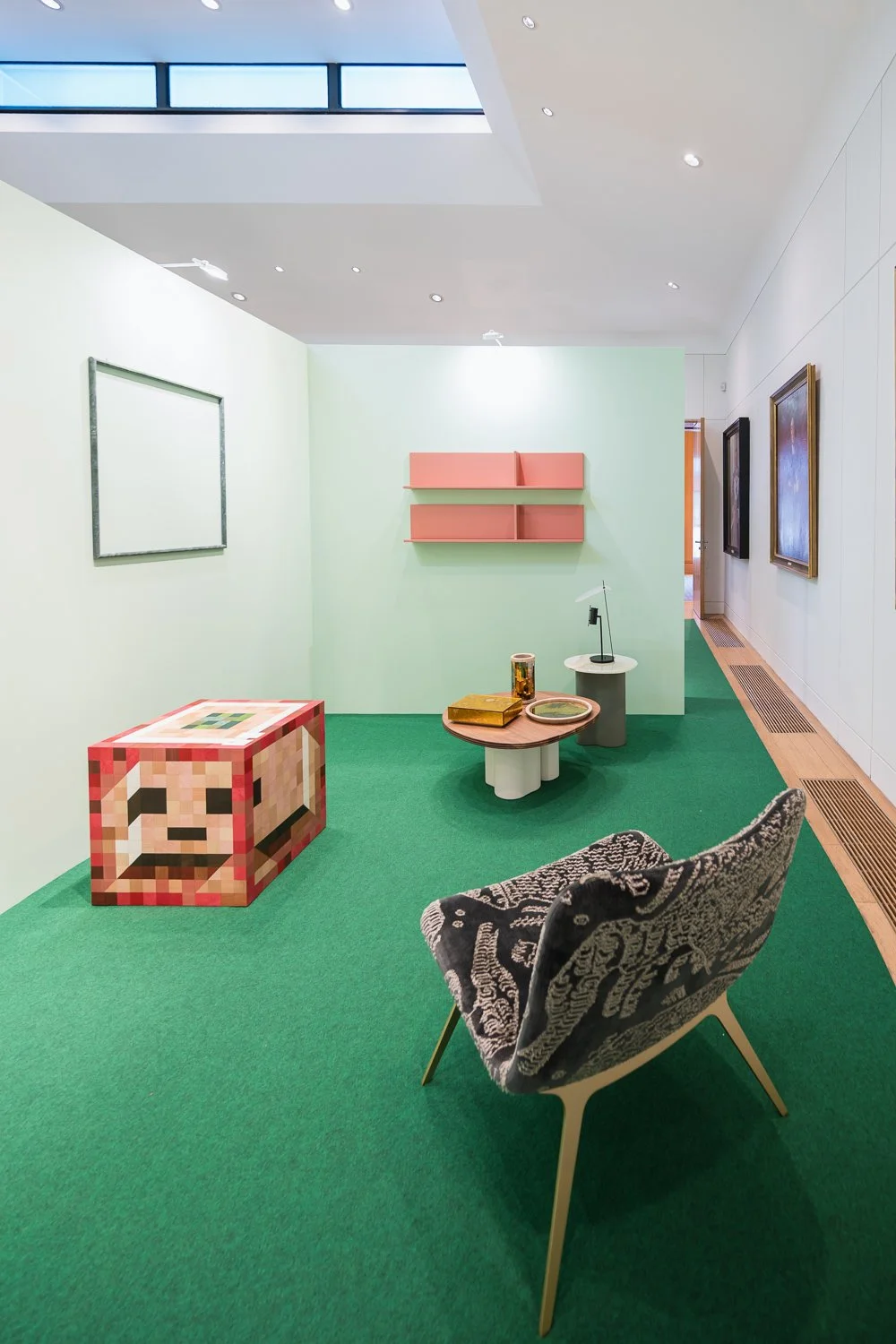 Art gallery room with green carpet, white walls, and colorful artwork, including a pixelated pixel art block, a pink floating shelf, and various framed paintings, with modern furniture like a patterned chair and circular tables.