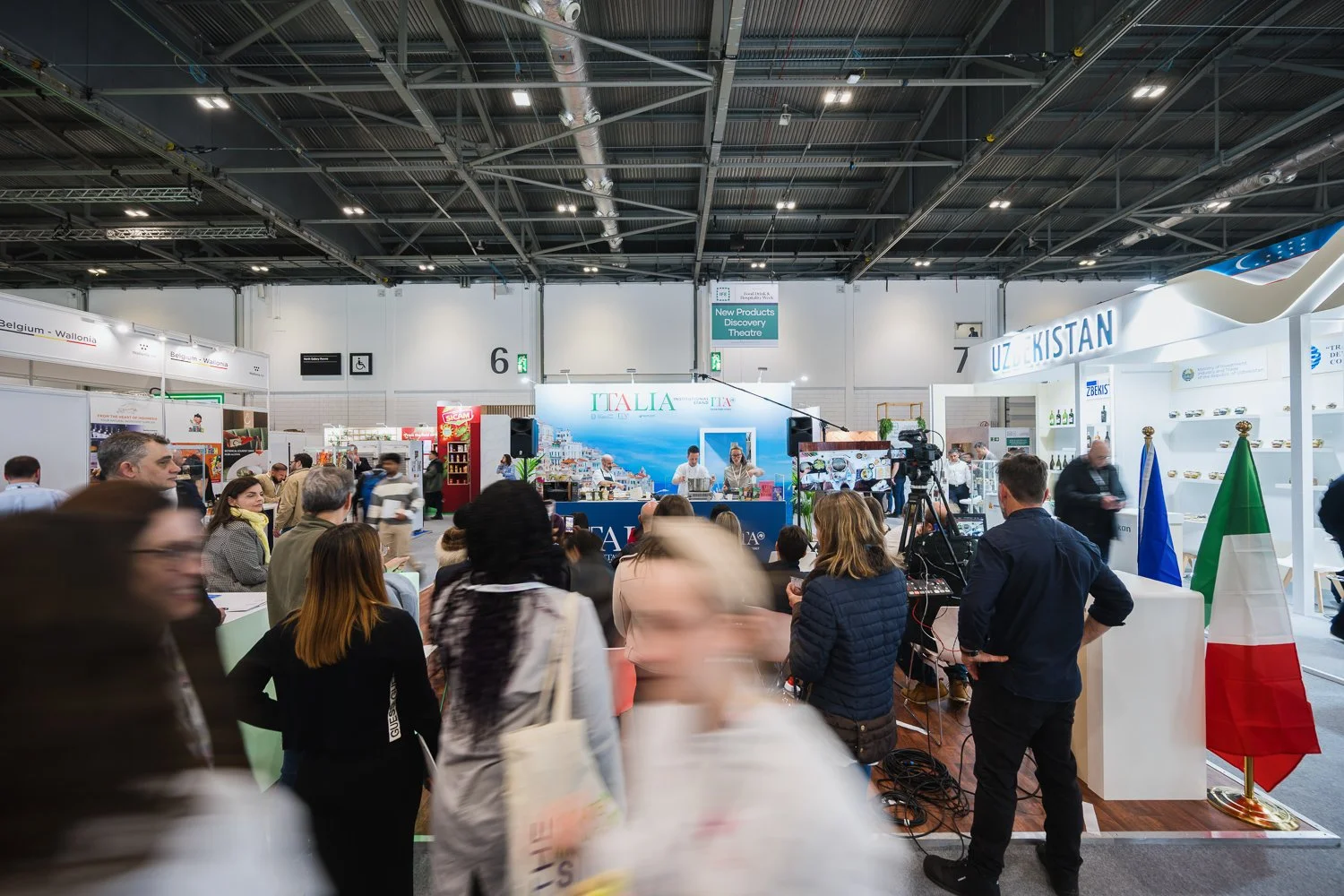 Crowd attending an exhibition or trade fair with booths lining the hall, including a central Italy booth with a scenic backdrop and a Uzbekistan booth with flags, and people engaging with displays and filming.
