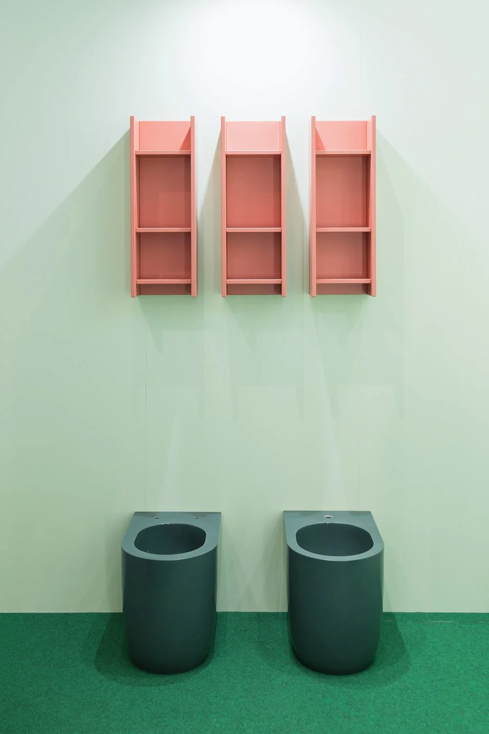 Contemporary bathroom with two black bidets, pink wall-mounted shelves, and a green carpeted floor.