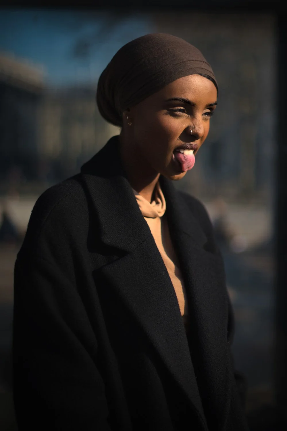 A young woman with dark skin, wearing a headscarf, sticking out her tongue playfully, dressed in a black coat outdoors.