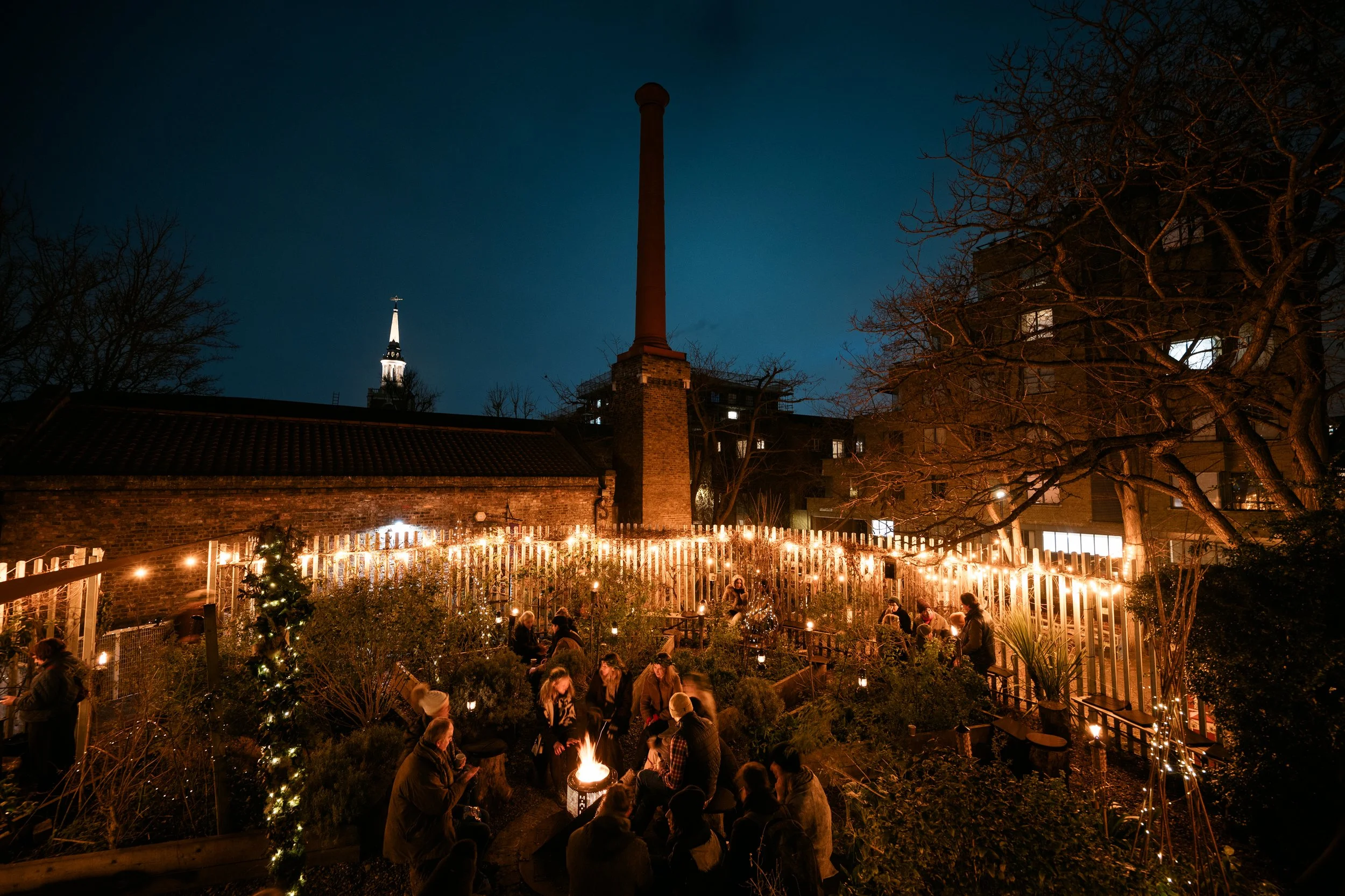 A nighttime scene of an outdoor gathering with people sitting around a fire pit, decorated with string lights and a small Christmas tree, set in a courtyard with brick buildings, leafless trees, and a view of a church steeple illuminated against the 