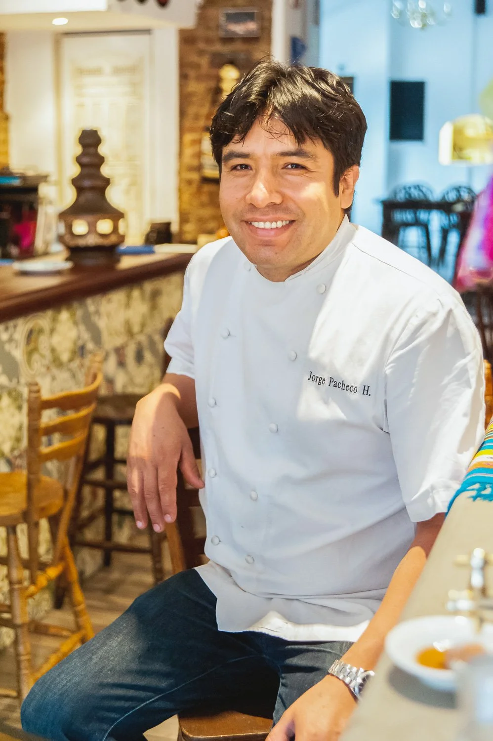 A smiling male chef wearing a white chef's coat with the name 'Jorge Pacheco H.' sitting in a restaurant setting with wooden chairs and decorative elements in the background.