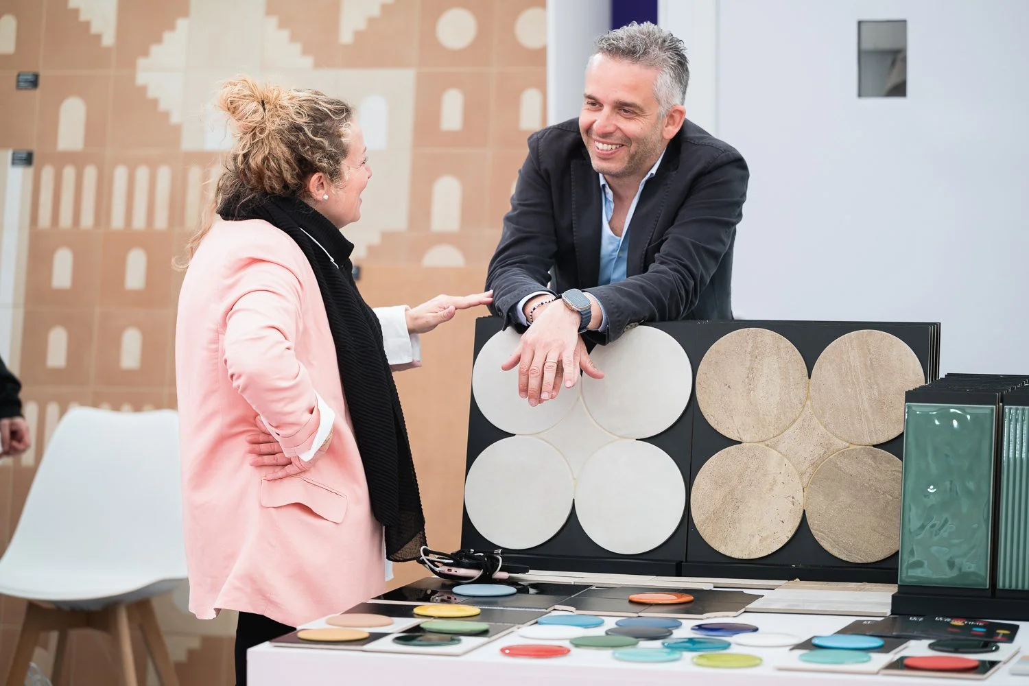 A woman in a pink blazer and black scarf engaged in conversation with a smiling man in a black blazer, who is leaning on display samples of various colored and textured material samples at a trade show or exhibition.