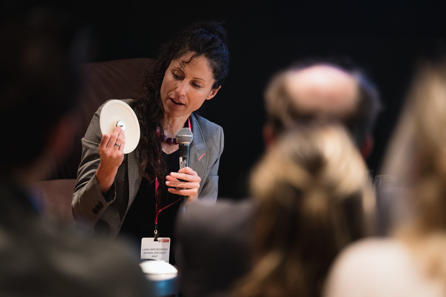 A woman speaking at a conference, holding a round object and a microphone, with a name tag reading "Laura Marcheggiano, Material Specialist". She has dark curly hair and is wearing a gray blazer.