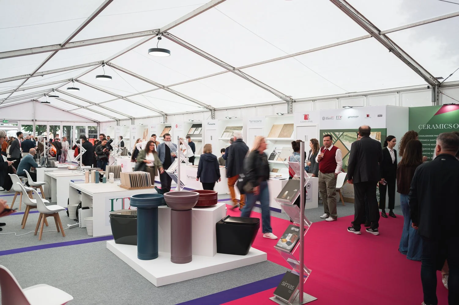 Trade show inside a large white tent with various booths displaying ceramics and home decor items. People are walking around, browsing, and talking to vendors.