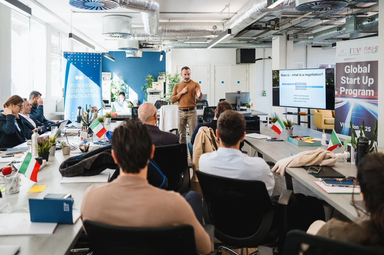 Business meeting or workshop in a modern conference room with a presenter standing near a large screen displaying a presentation, surrounded by seated participants with laptops and Italian flags on the table, and banners promoting corporate innovatio