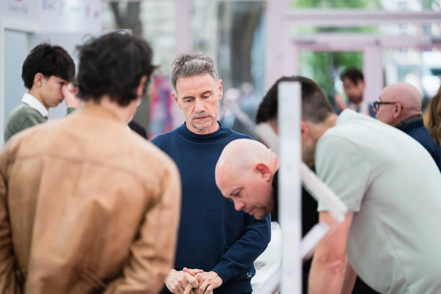 A group of people gathered around a counter in a store, with a man in a navy blue sweater at the center, looking down with a somber expression. The others are focused on the counter or engaged in activities, in a well-lit, modern retail environment.
