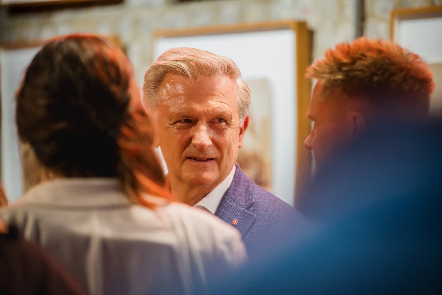 A middle-aged man with gray hair talking to two younger people at an art gallery or similar setting.