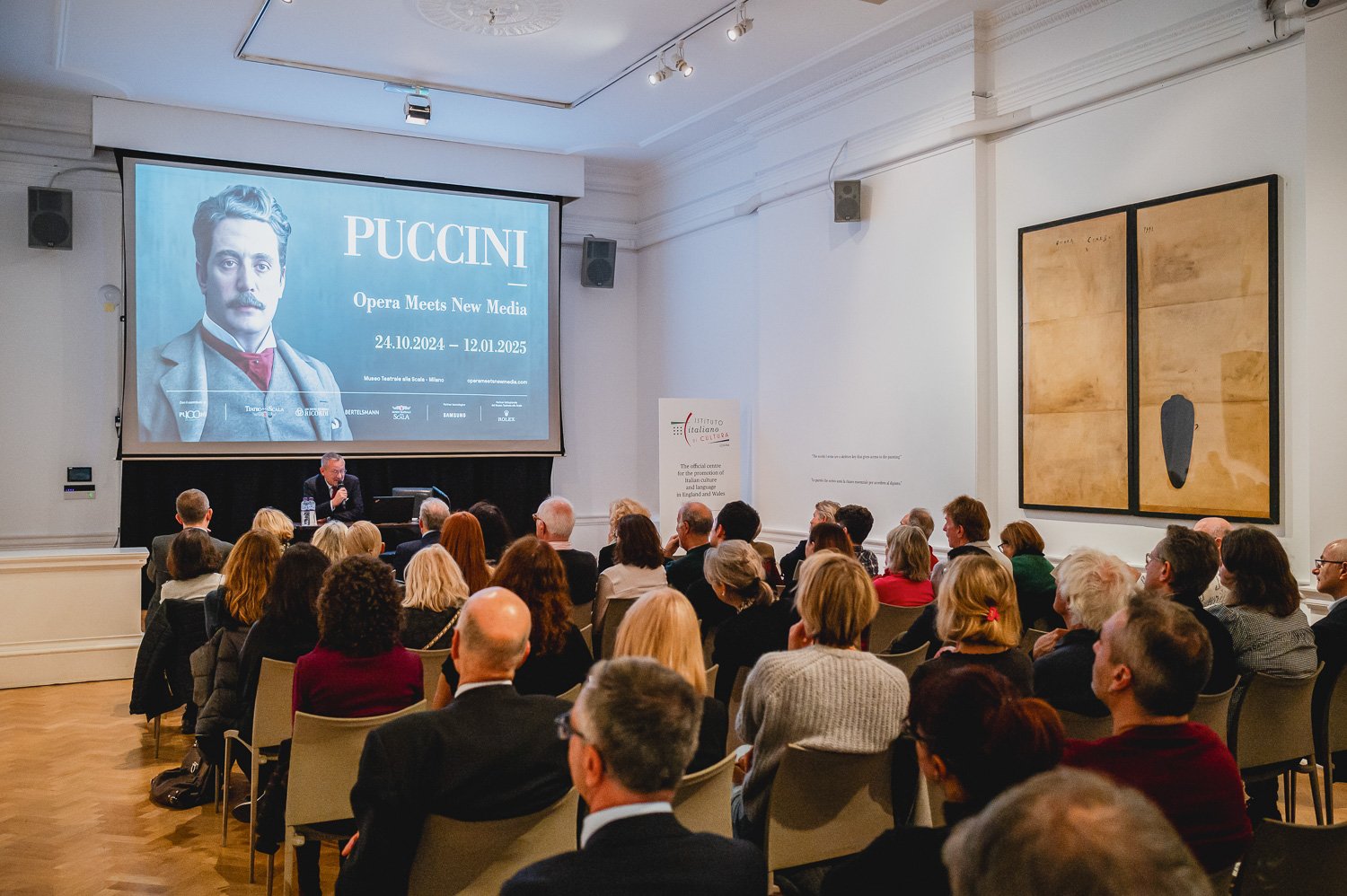 A conference room filled with people attending a presentation. A man is speaking at a desk near the front, with a large screen behind him displaying text about Puccini and opera meeting new media from October 24, 2024, to January 12, 2025. The room h