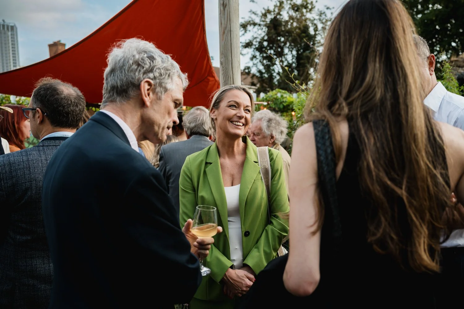 Group of people socializing outdoors at a gathering, including a smiling woman in a green blazer holding a glass of white wine, and others engaged in conversation.