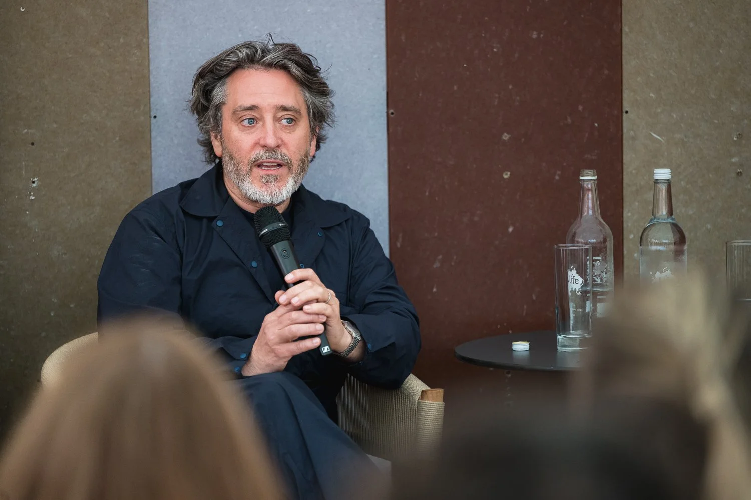 A man with gray hair and beard, wearing a black shirt, speaking into a microphone during a panel discussion, sitting at a table with bottles of water and glasses, with audience members in the foreground.