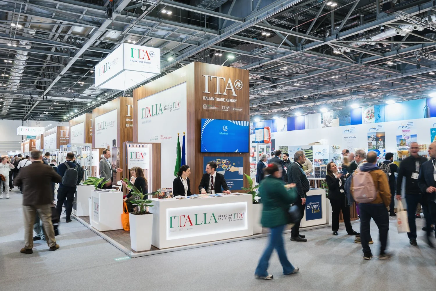 Trade exhibition hall with numerous booths, prominently featuring the Italian Trade Agency's stands with signage and branding, people engaging in conversations and exploring displays.