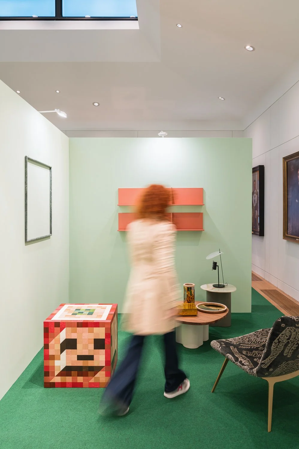 An art gallery room with a blurred person walking past colorful modern artworks and furniture, including a pixelated sculpture, framed paintings, and decorative tables on green carpet.