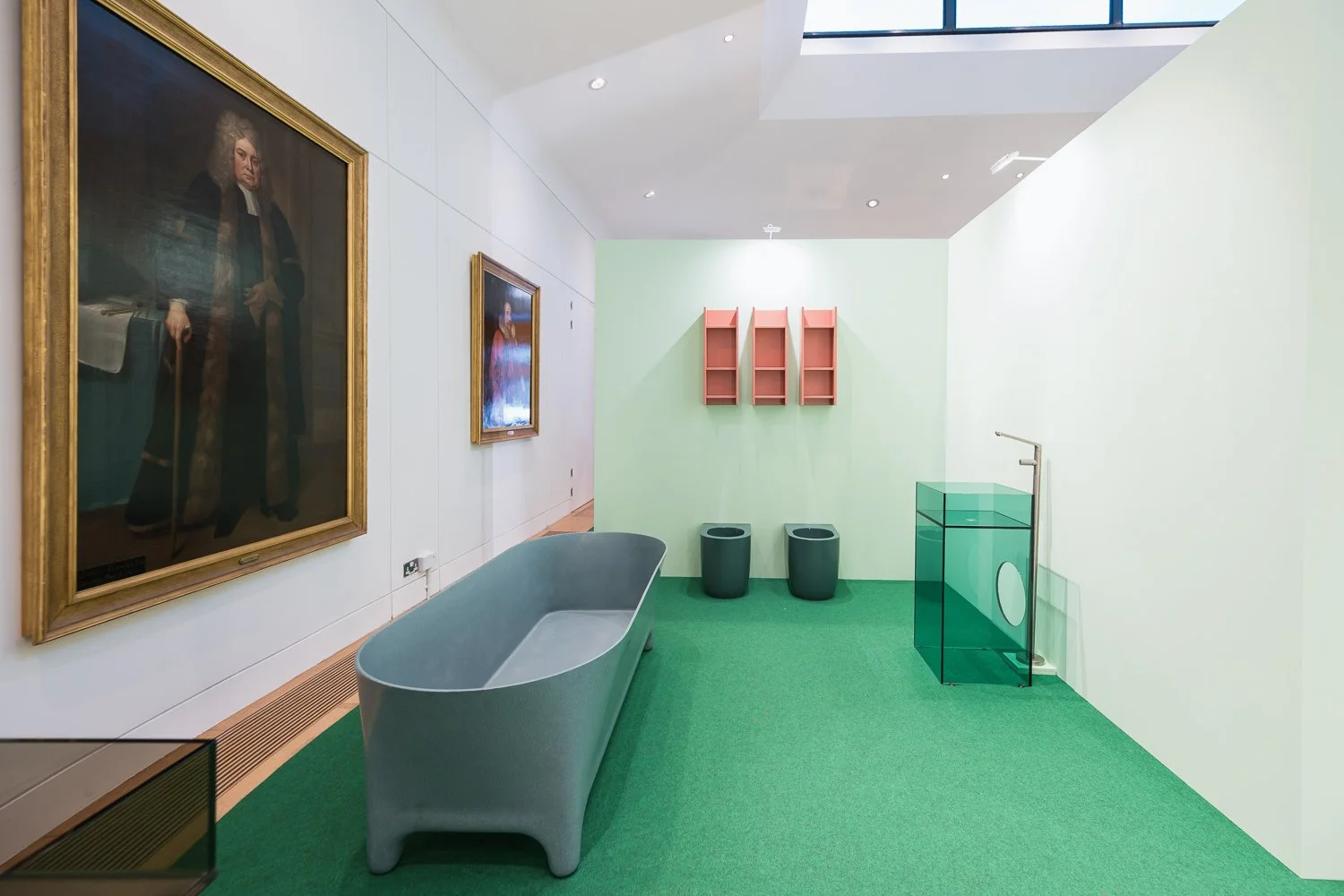 An art gallery with classical paintings on the white walls, a modern gray bathtub, pink wall shelves, two black trash bins, a transparent green display case, and a green carpeted floor, illuminated by natural light from a skylight.
