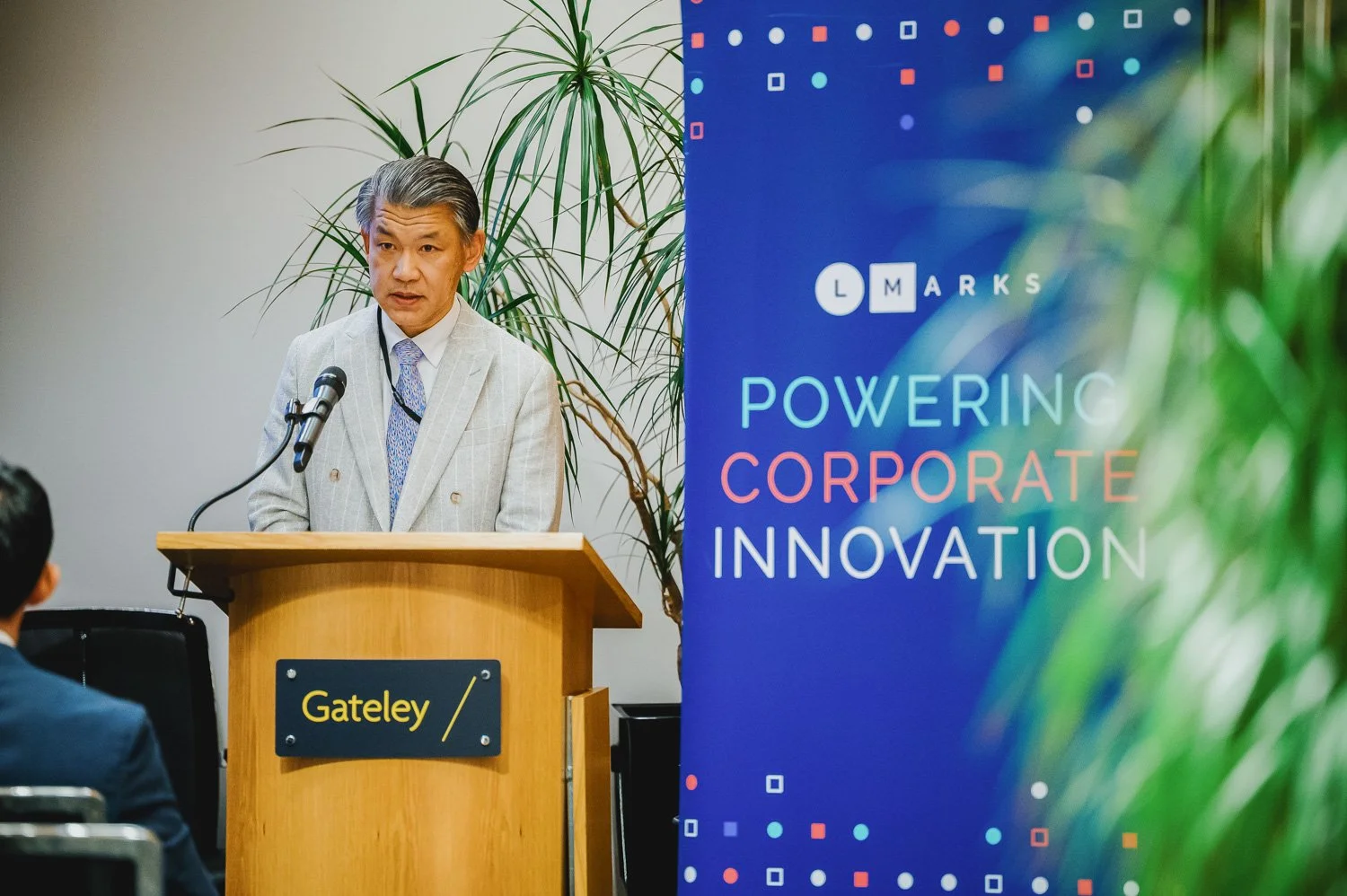 A man in a light gray suit and tie standing behind a wooden podium labeled "Gateley" speaking into a microphone during a corporate event. Behind him, a large blue vertical banner displays the words "Powering Corporate Innovation" with a colorful digi
