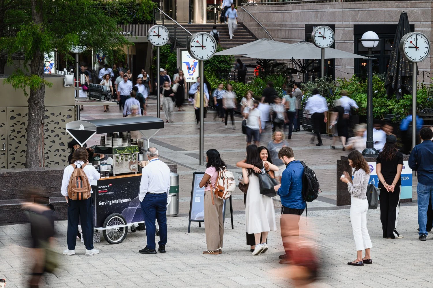 Crowded outdoor urban area with people walking, standing, and chatting. There is a coffee cart, large clocks on poles, greenery, stairs, a pizza stand, and signs for services.