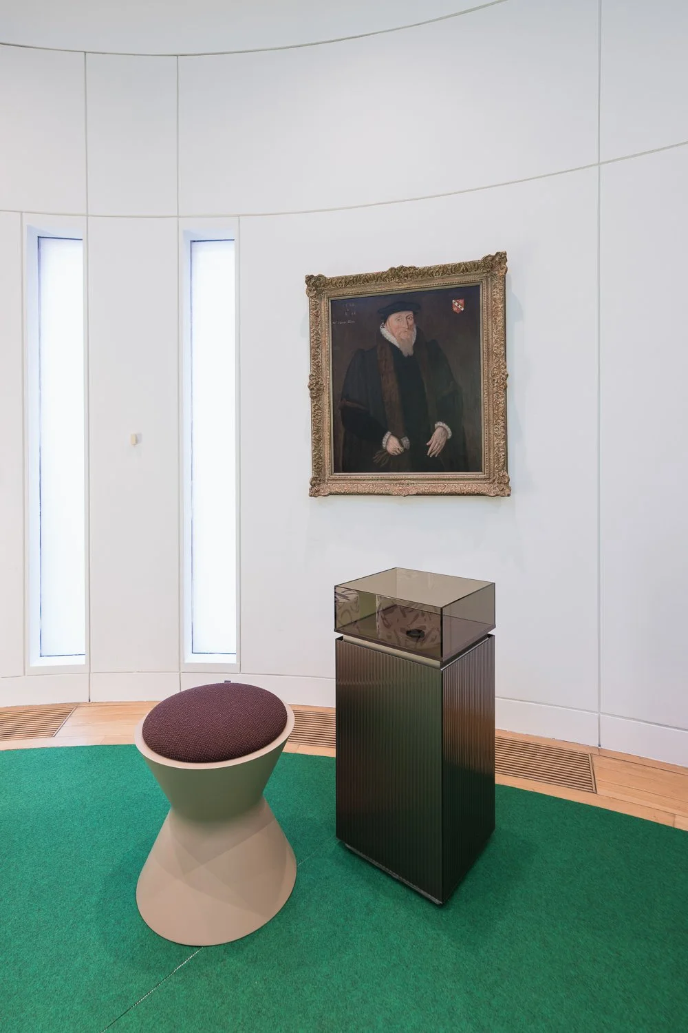 Modern art gallery interior with a framed portrait of a man in historical clothing on a white curved wall, a glass display case on a black pedestal, a contemporary round stool, and a green carpeted floor.