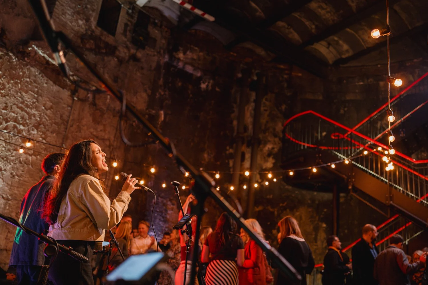 A woman singing into a microphone at a live music event in an indoor venue with an exposed brick wall and string lights, surrounded by an audience.