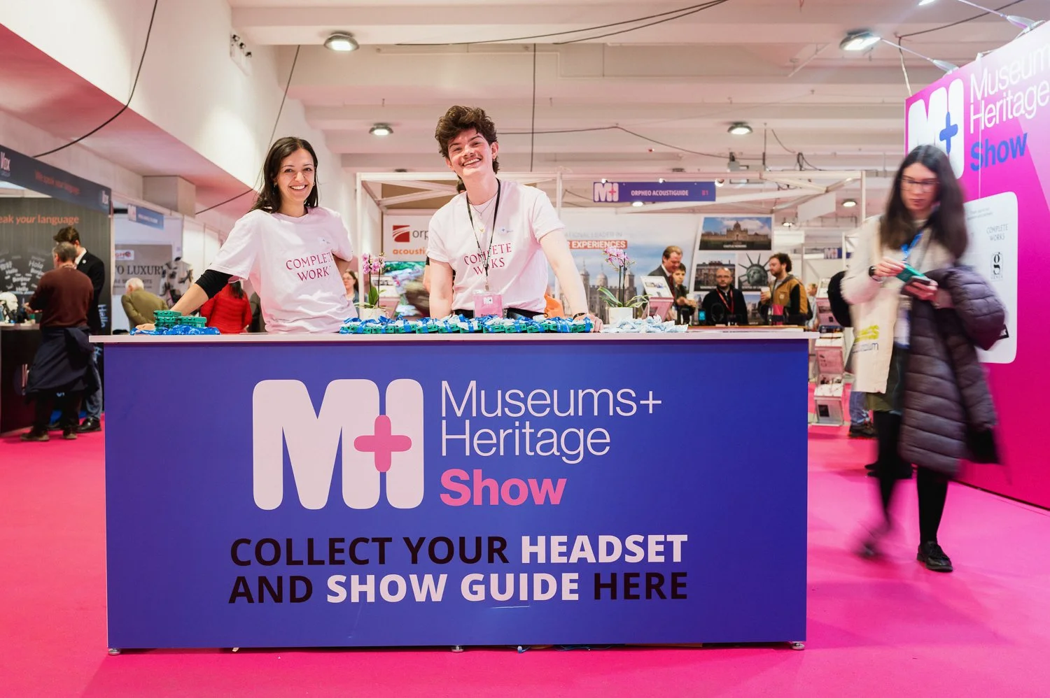 Two smiling women standing behind a registration desk at the Museums + Heritage Show, with a woman walking by on the right. The desk has a sign that reads 'Collect your headset and show guide here.' The background shows other booths and attendees at 