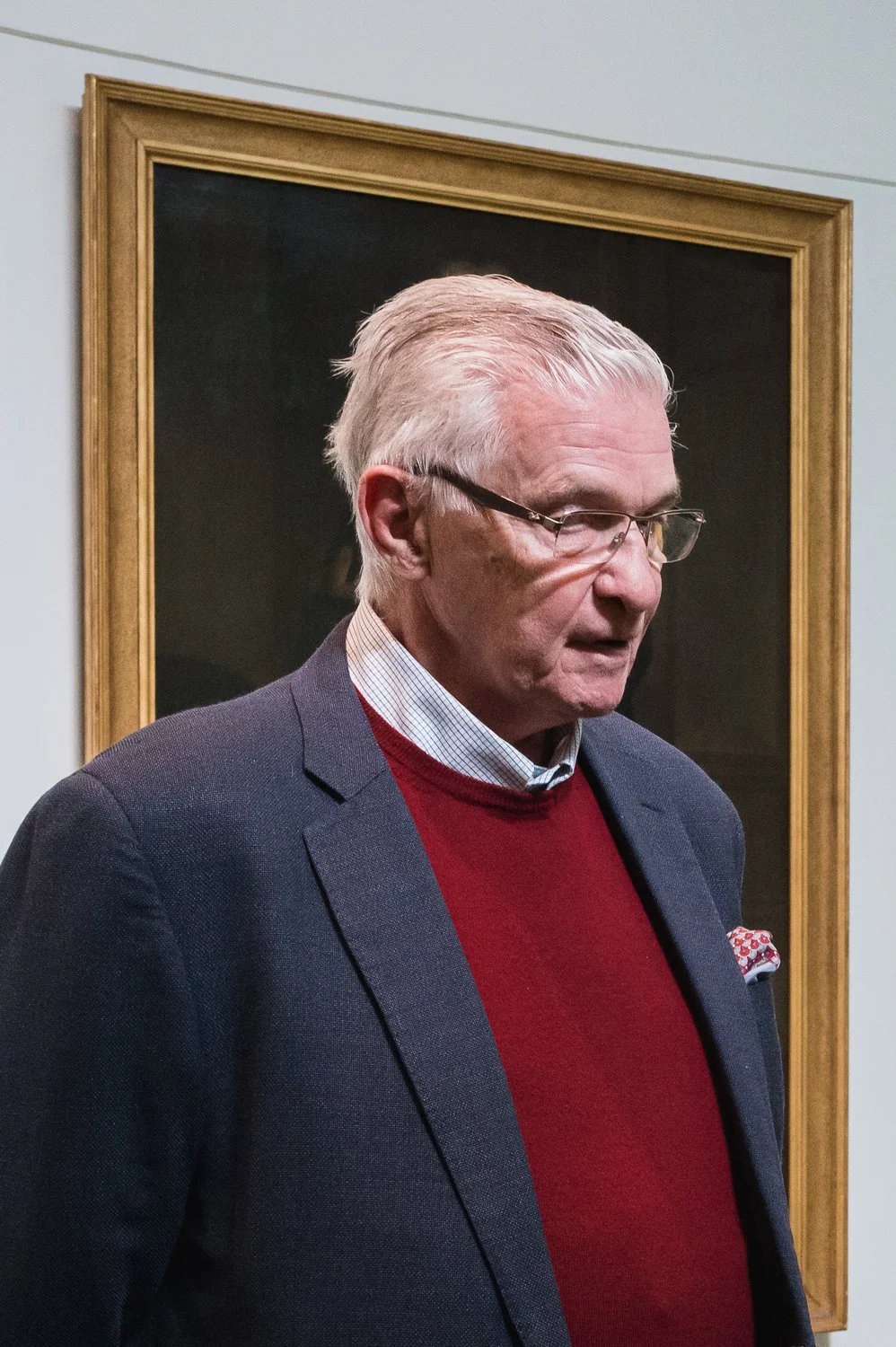 An older man with glasses and white hair wearing a navy blazer, red sweater, and collared shirt, standing in front of a framed portrait painting.