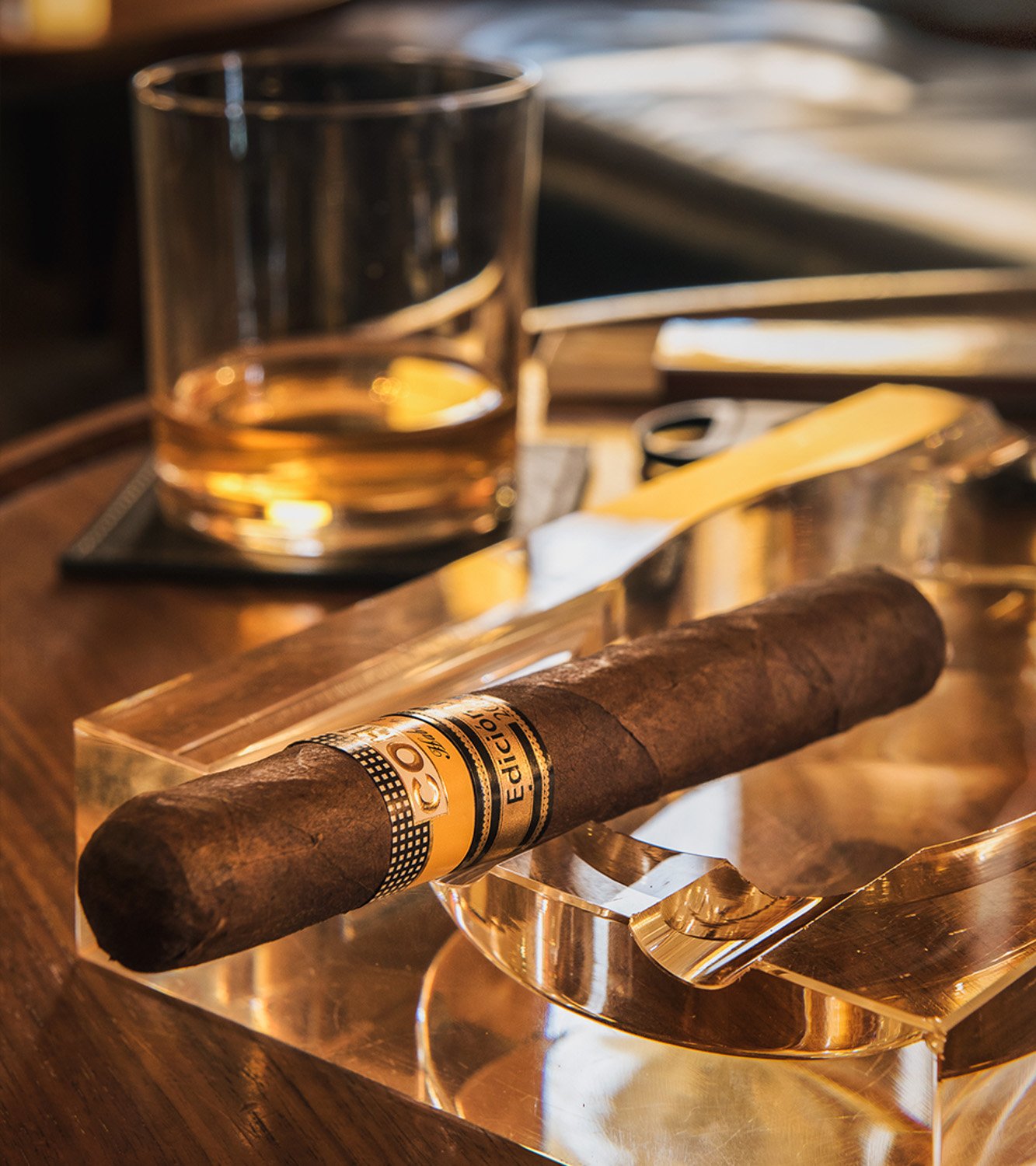 A cigar resting on a glass ashtray with a glass of whiskey, a lighter, and papers on a wooden table.