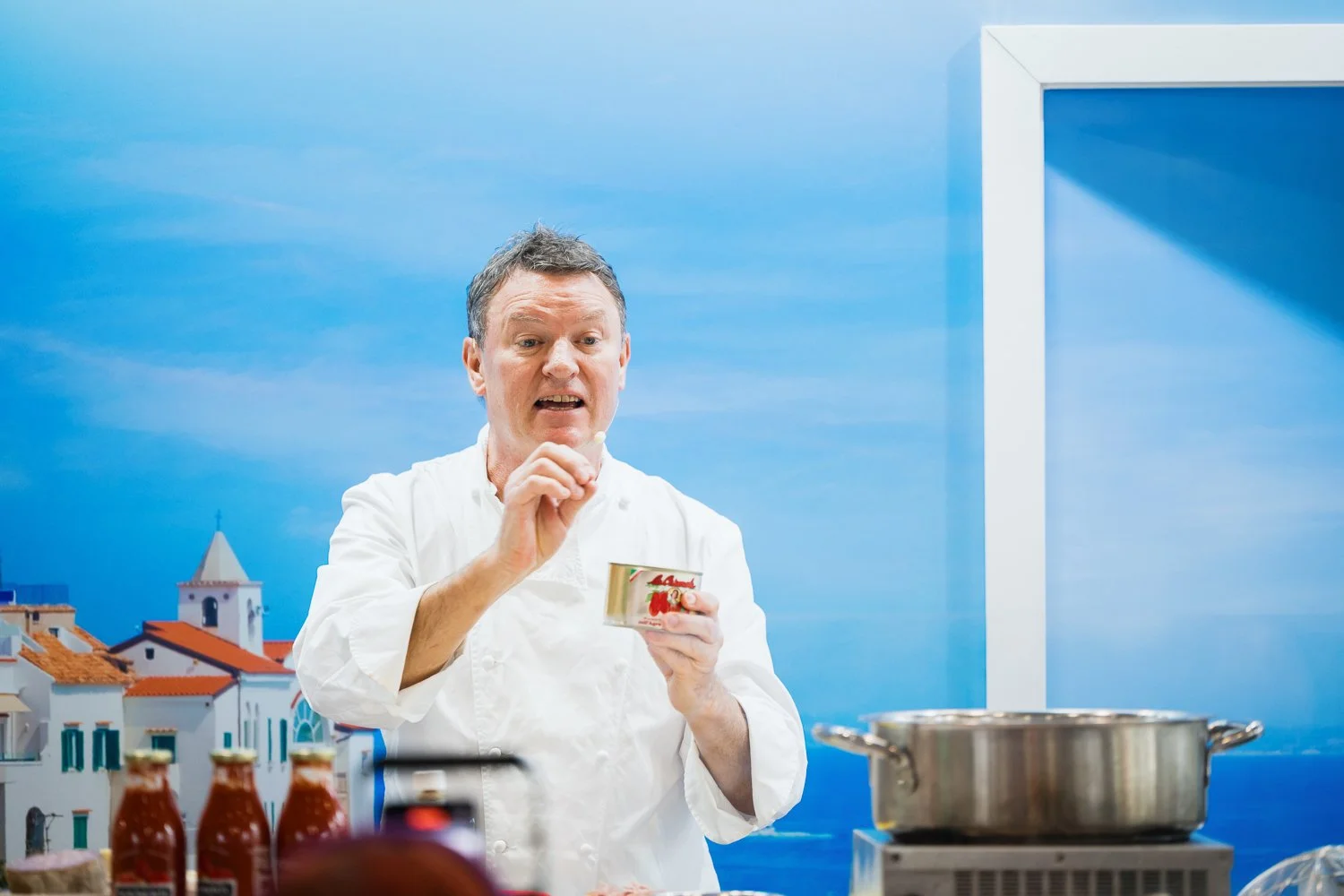 Chef in a white uniform holding a can of tomatoes, speaking in a kitchen with a blue sky and coastal village background, with bottles of sauce and a pot on the stove in the foreground.