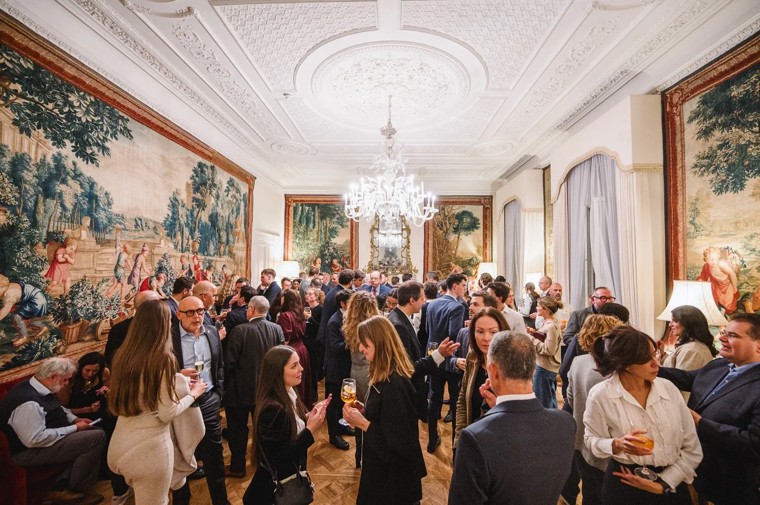 An ornate room filled with people attending a social gathering or party, with large tapestries on the walls, a chandelier hanging from the ceiling, and elegant lighting.