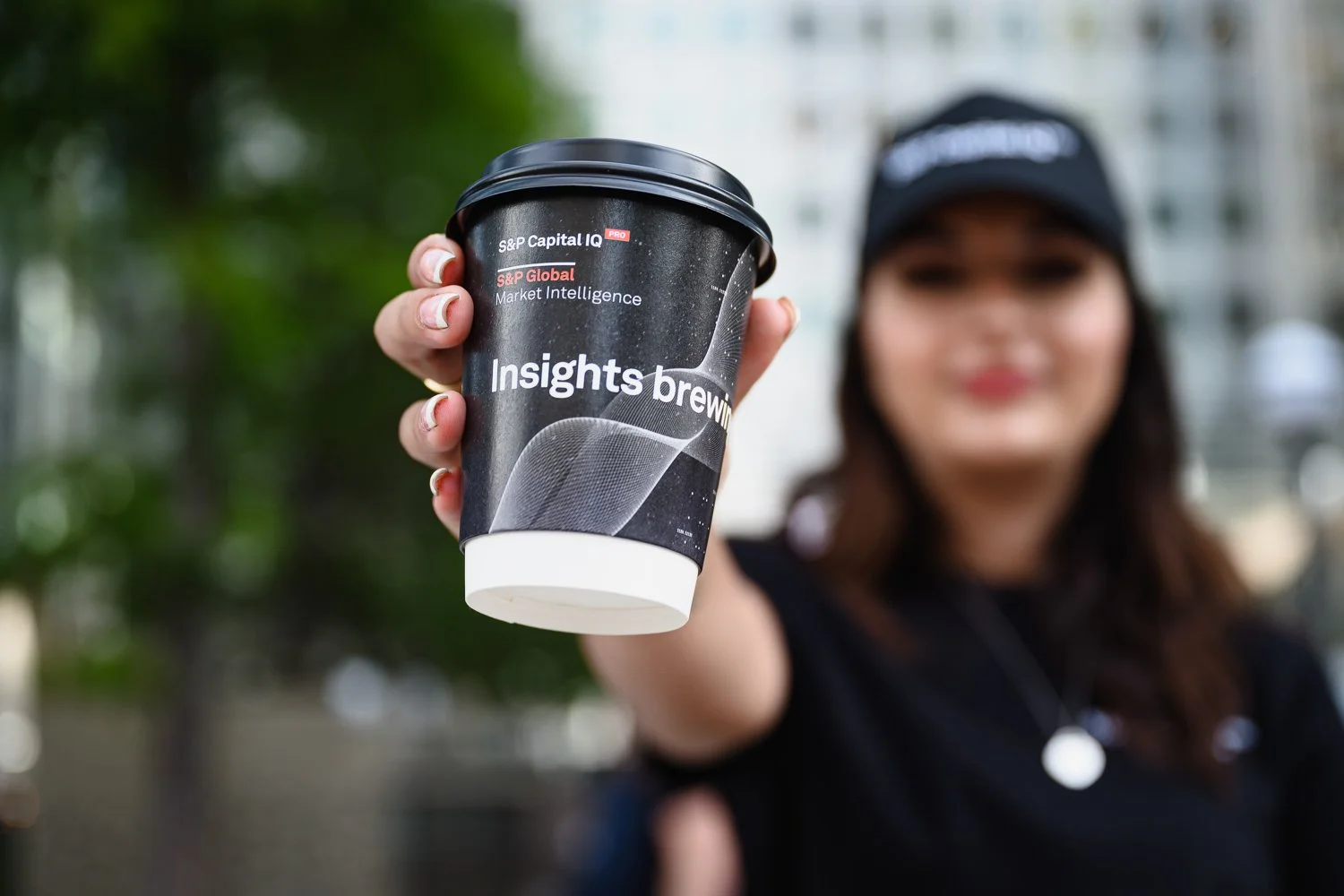 A woman holding a black coffee cup with the logo 'Insights Brewing' towards the camera, outdoors with blurred trees and buildings in the background.