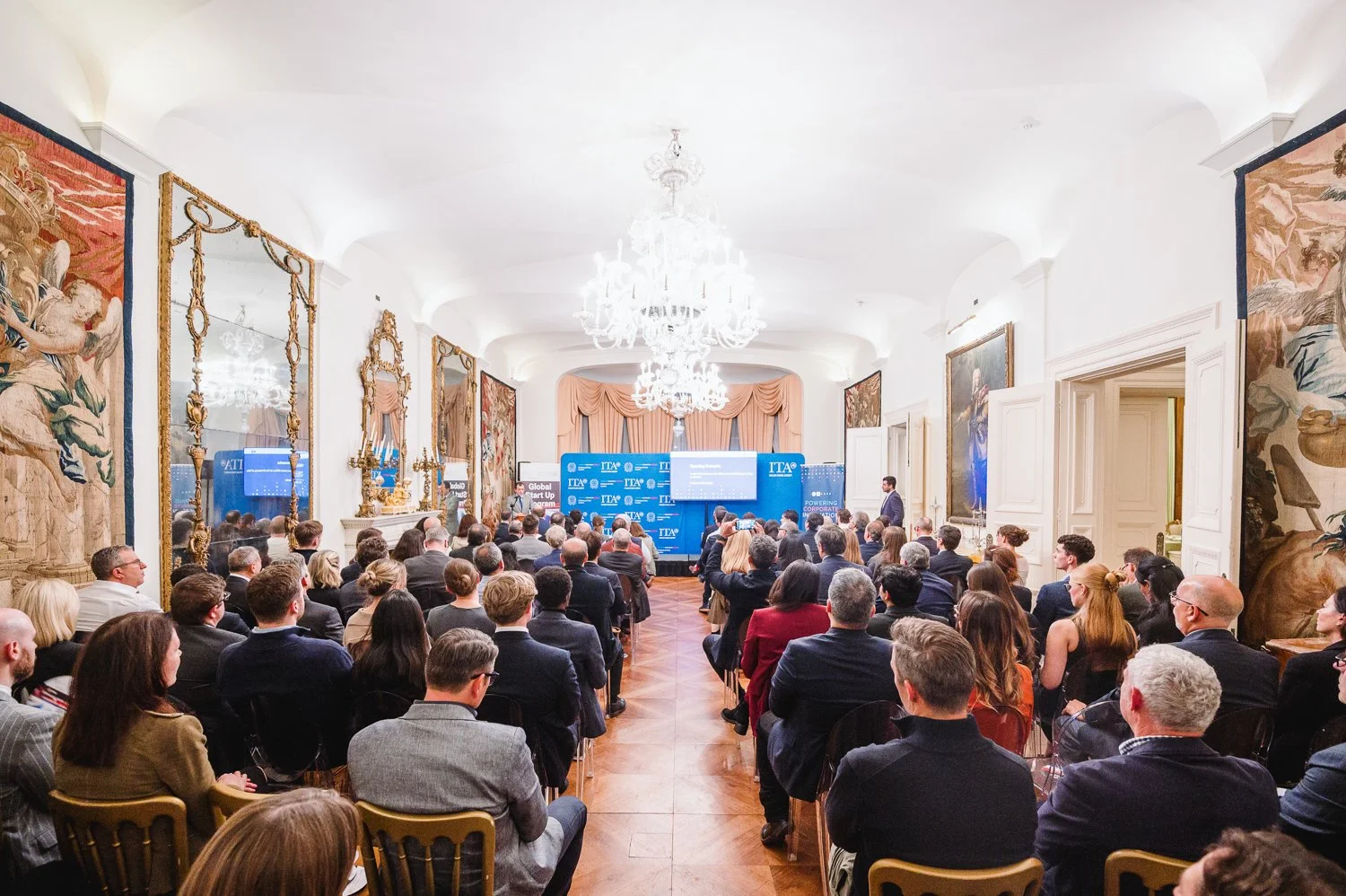 A conference or formal presentation event is taking place in an elegant room with high ceilings, ornate wall decorations, and a large chandelier. The room is filled with seated attendees facing a stage with speakers and presentation screens.