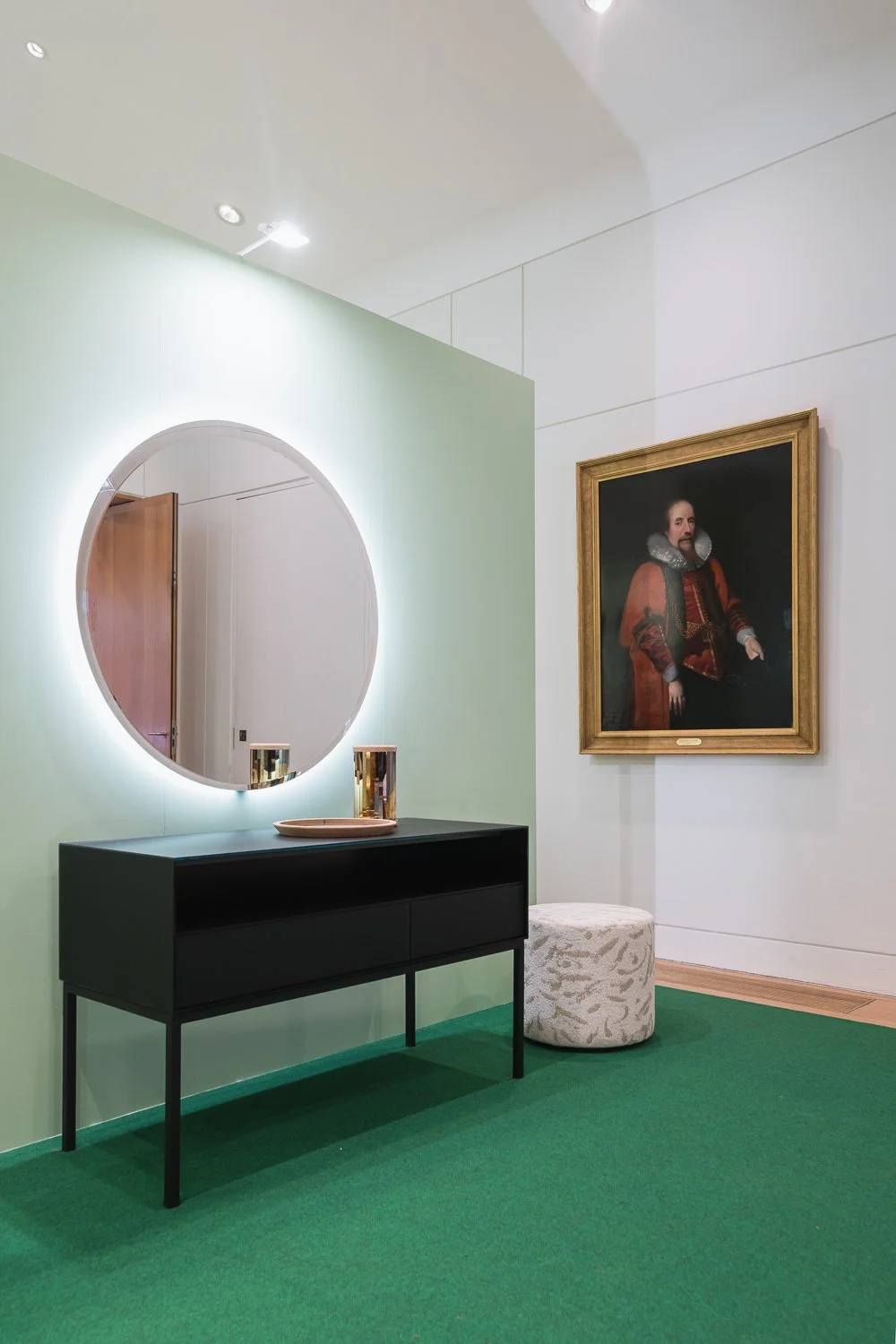 A modern interior space with a black console table, a round mirror with backlighting, and decorative items on top. To the right, there is a framed portrait of a man in historical attire, and a textured round ottoman beside it. The floor is green carp