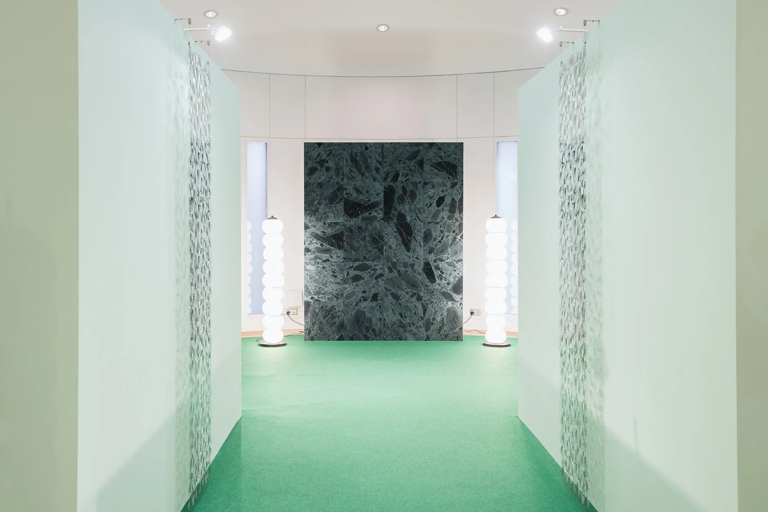 A modern interior space with a green carpet, white walls, and a black marble wall in the background. Two tall, white, spherical floor lamps are on either side of the marble wall. Vertical windows on either side of the marble wall allow natural light 