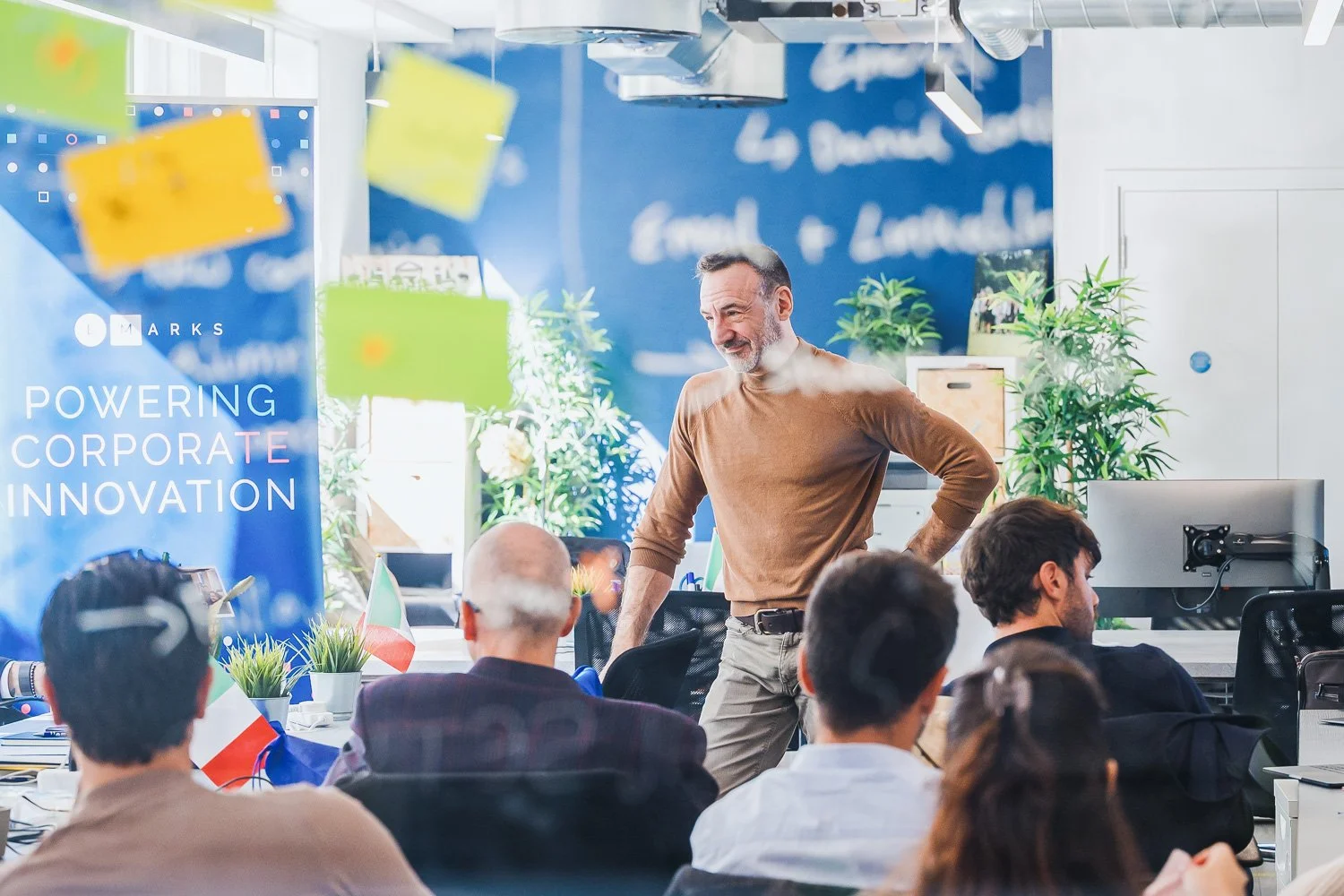 A man with a beard and wearing a brown shirt stands in a bright office, smiling and talking to a group of people seated at a table. The office has colorful sticky notes hanging in the air and a blue wall with white text that reads 'POWERING CORPORATE