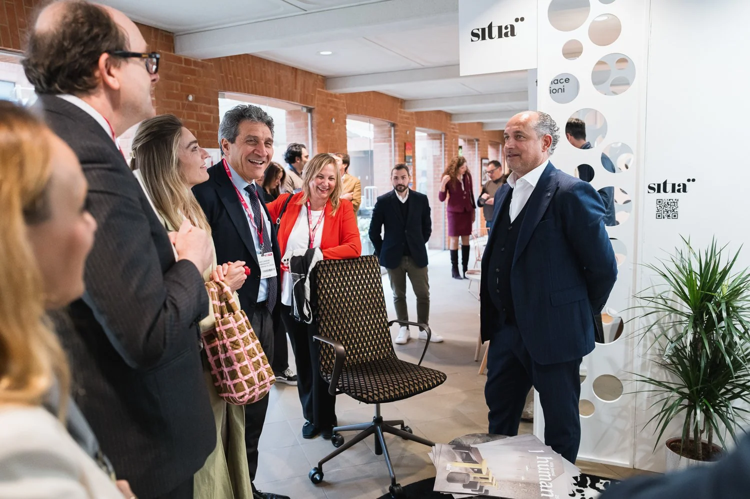 A group of business professionals, including men and women, engaging in a conversation at a conference or event. They are smiling and appear to be enjoying the interaction. The setting is a modern indoor space with brick walls, large windows, and a w