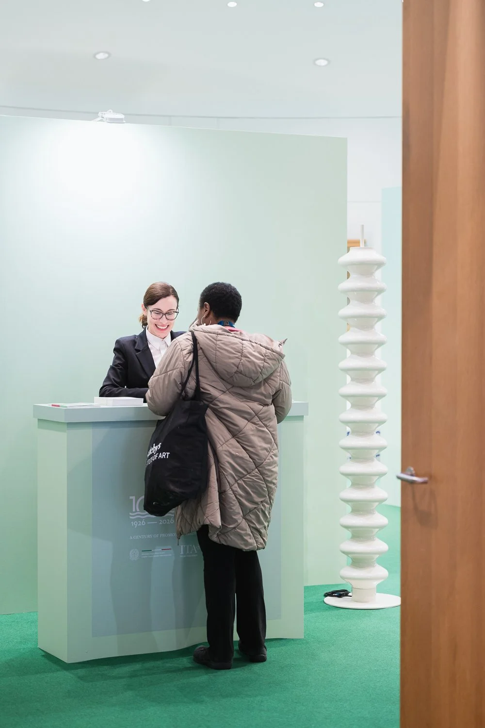 A woman at a reception desk talking to a guest in an exhibition or event space with green walls and a unique white sculpture on the right.