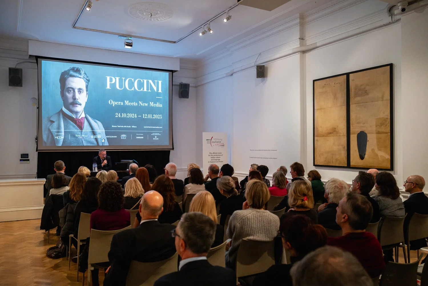A conference room filled with seated attendees watching a presentation. A man is speaking at a podium in front of a large screen displaying a promotional image for an event about Puccini, titled "Opera Meets New Media," held from October 24, 2024, to