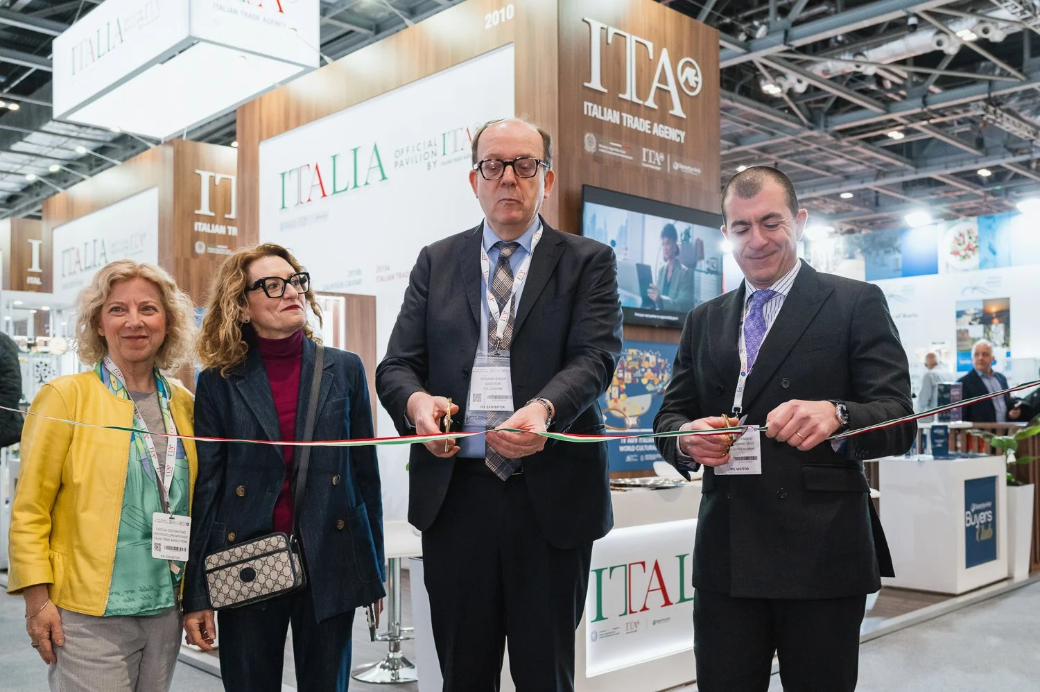Five individuals participating in a ribbon-cutting ceremony at the Italian Trade Agency booth. Two men are cutting the ribbon, while three women watch smiling. Booth signage includes 'ITALIA' and 'ITALIAN TRADE AGENCY'.