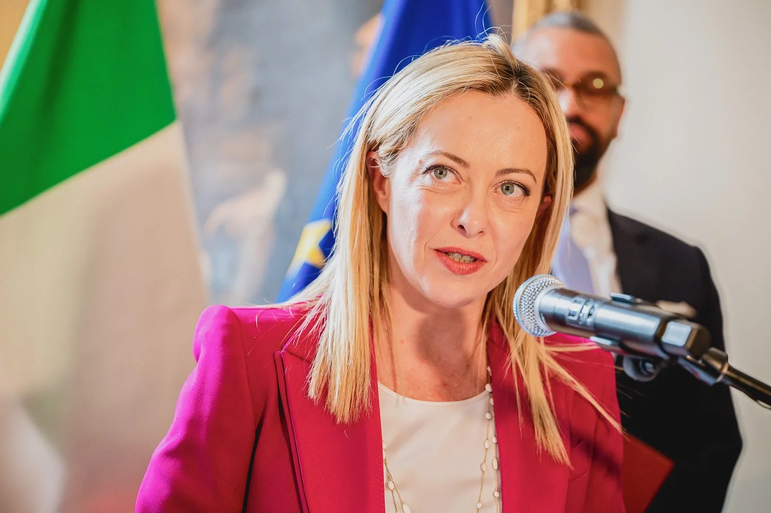 Giorgia Meloni wearing a red blazer speaking into a microphone at a press conference, with a man in a suit and glasses in the background and flags visible behind them. Phocus Collective Event Photography.