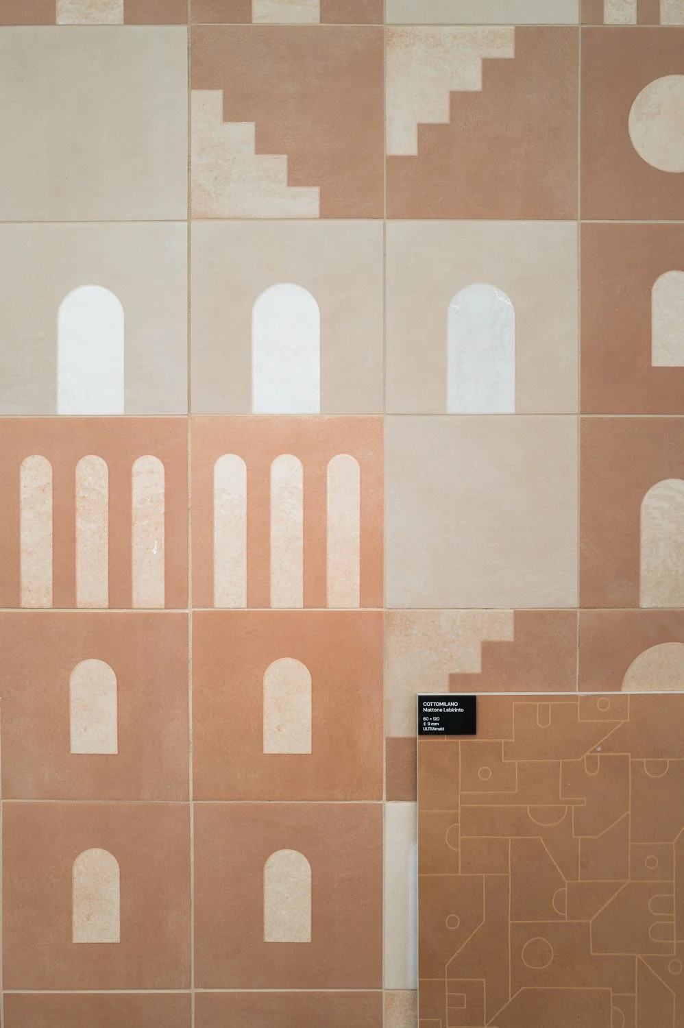A wall decorated with multiple square tiles, each with geometric patterns and varying shades of beige and terracotta. Some tiles feature stair-step and arch patterns, creating a modern, abstract design.