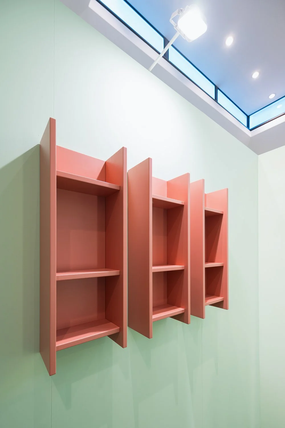 Pink wall-mounted shelves with no items on them, against a light green wall, with a ceiling light and skylight above.