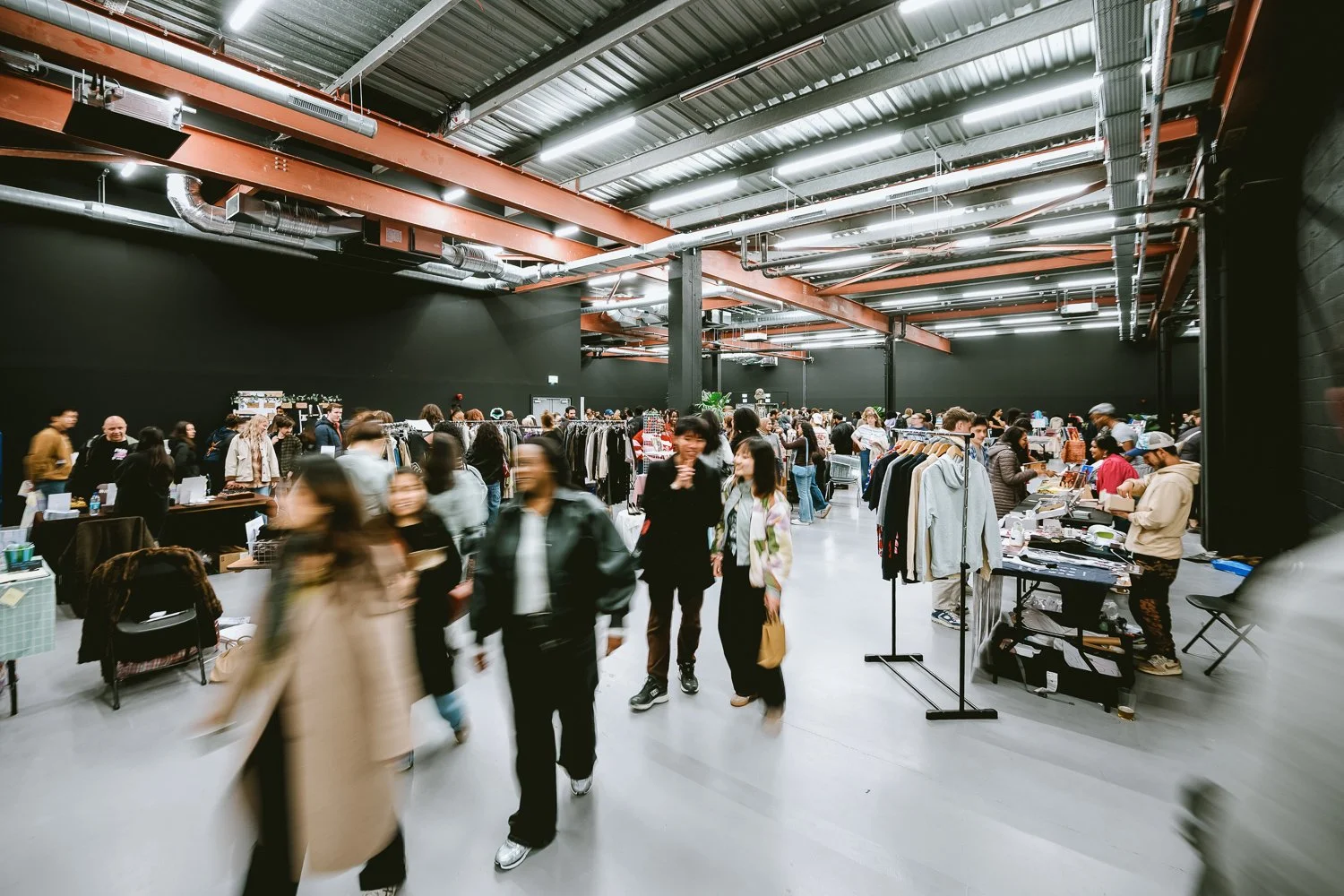 People shopping at a thrift store or flea market with racks of clothes and tables with various items, in a large industrial-style space with black walls and exposed ceiling beams.