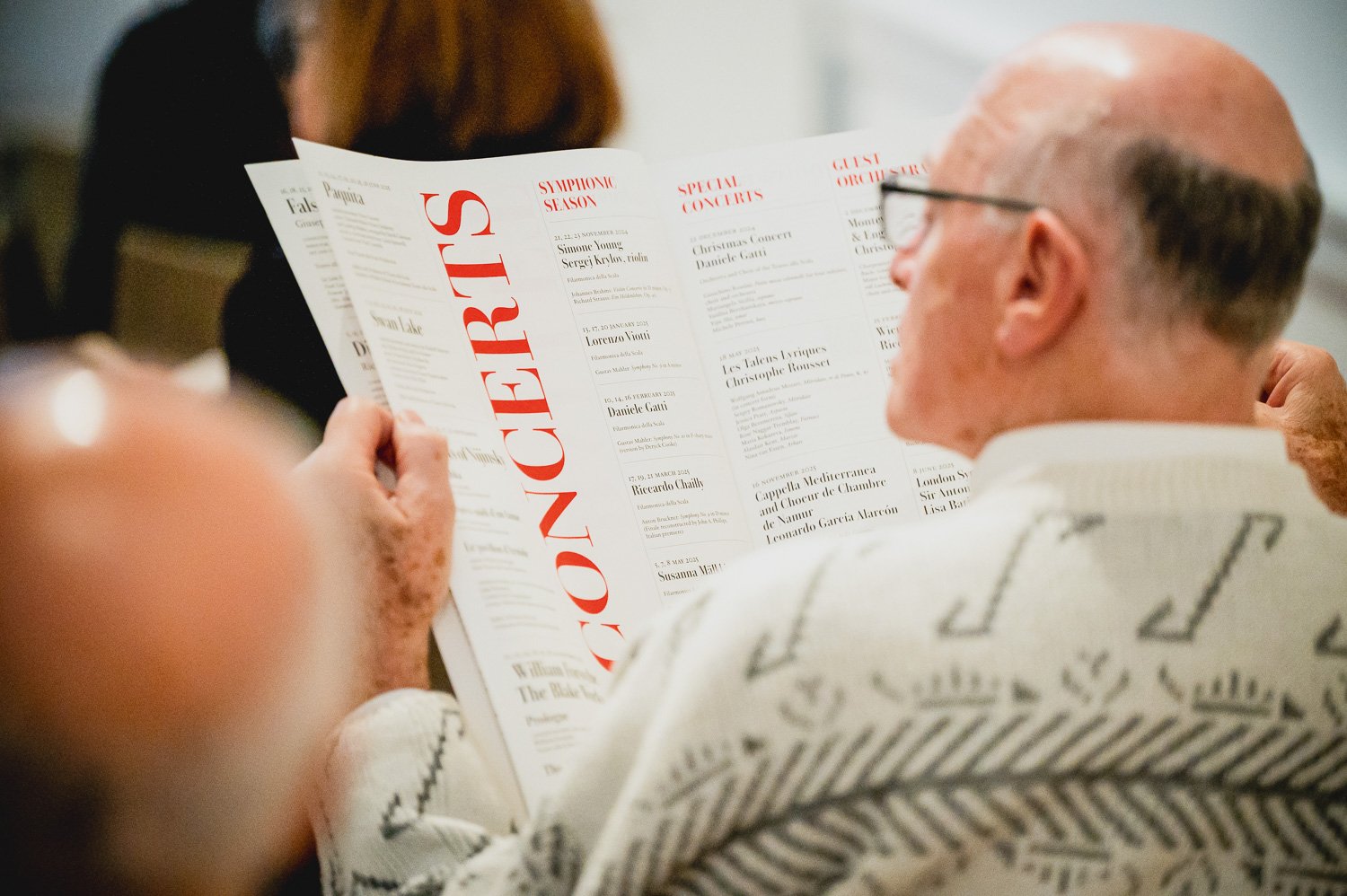 A man with glasses reading a program or flyer at an event, with a person in front holding a similar document, in an indoor setting. Phocus Collective Event Photography.