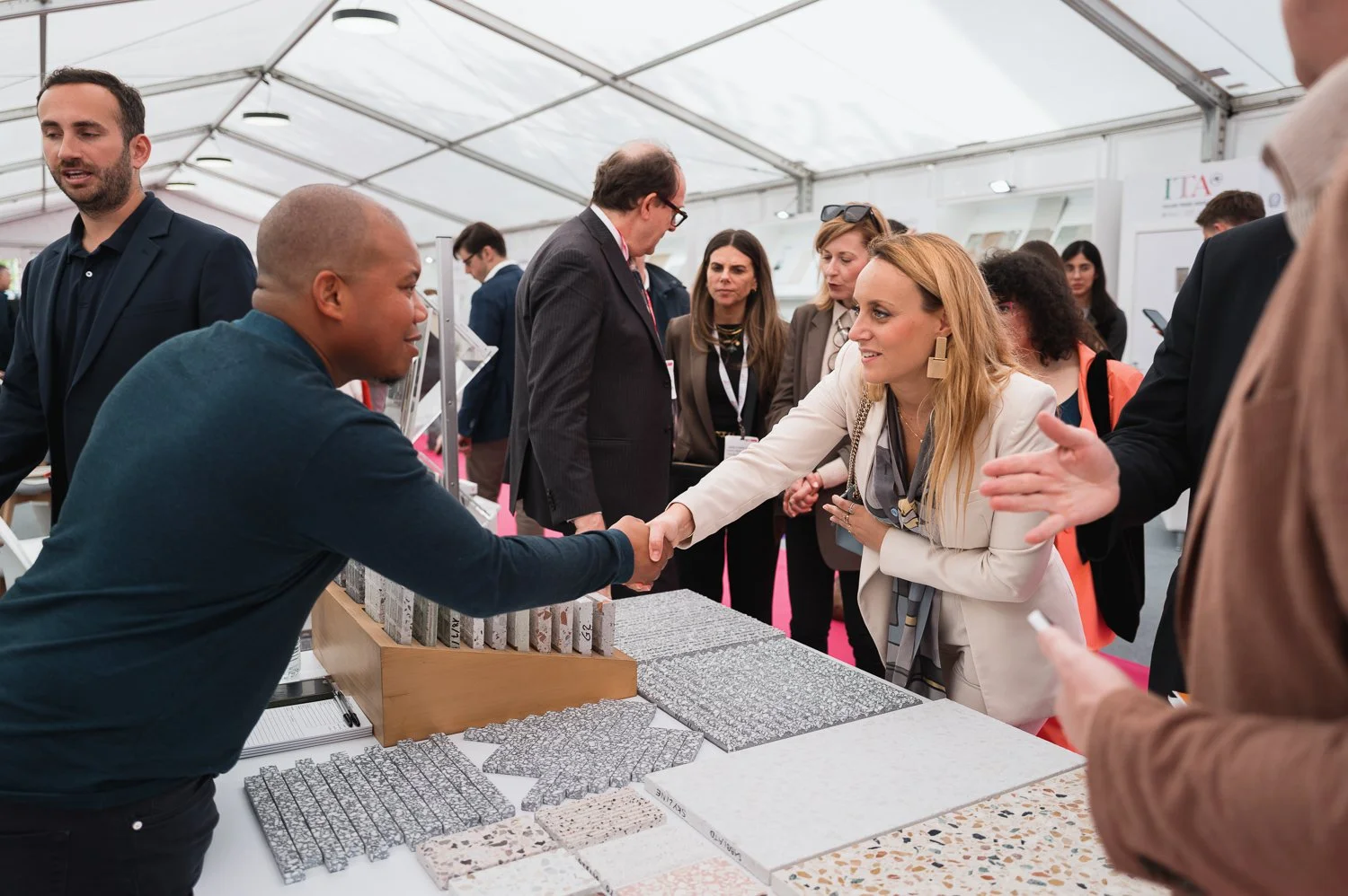 Businesswoman shaking hands with a man at a trade show booth displaying tile samples, while other people engage in conversations around.