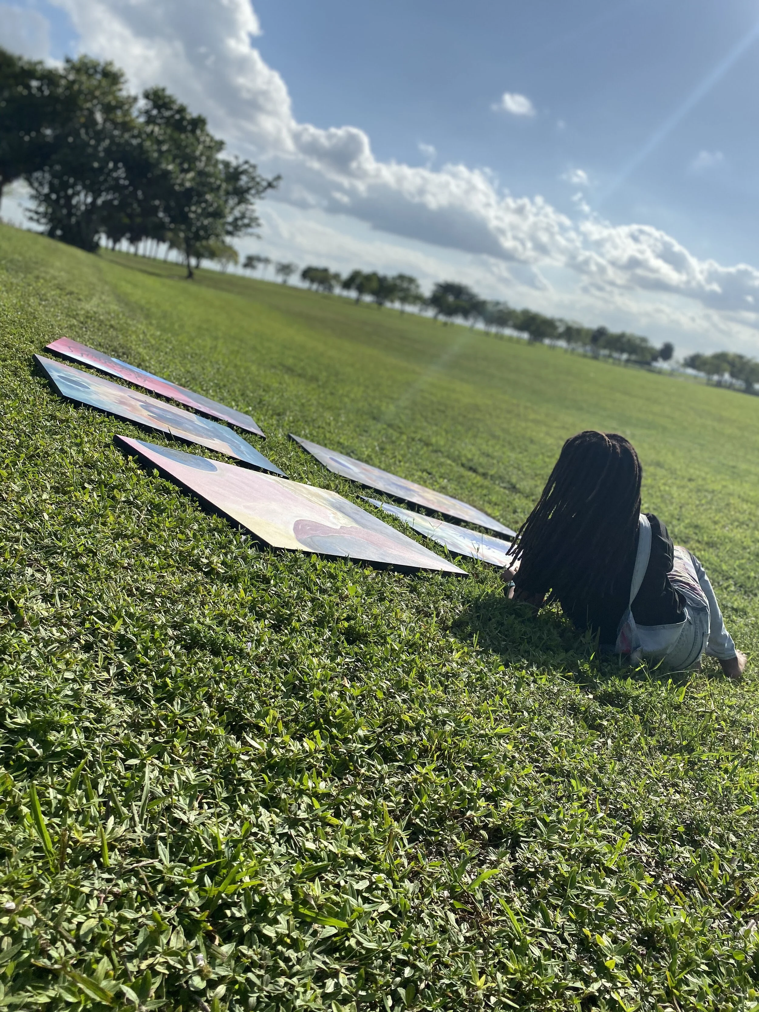 A person with long locked hair sitting on green grass outdoors, looking away from the camera, with large colorful artwork lying on the grass nearby, under a bright blue sky with scattered white clouds and trees in the background.