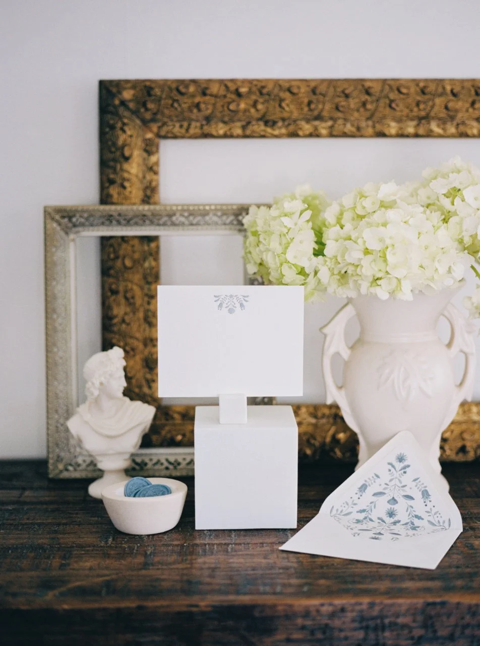 Elegant wedding stationery display with white cards and envelopes featuring blue floral designs, surrounded by white hydrangeas in a vase, ornate picture frames, a small bust statue, and wax seals in a bowl on a wooden table.