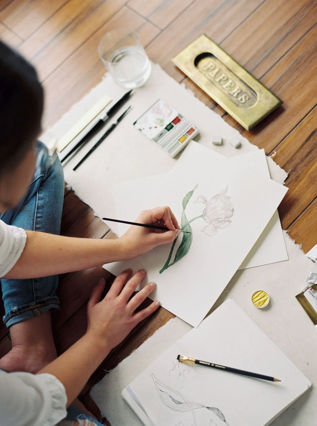 Person painting a flower on paper with watercolor paints and brushes on a wooden floor.
