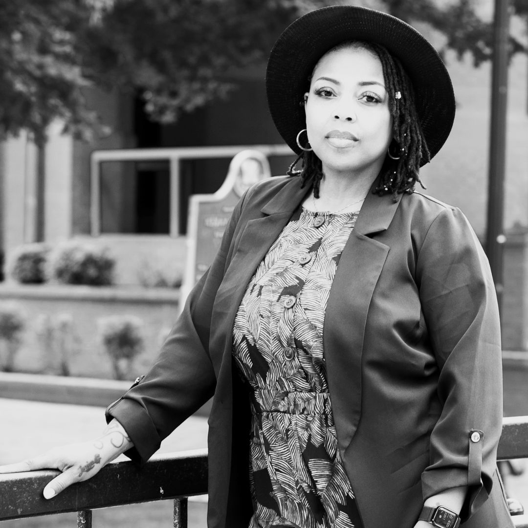 Black and white photo of a woman wearing a large hat, hoop earrings, and a patterned blouse, standing outdoors with her hand resting on a railing.