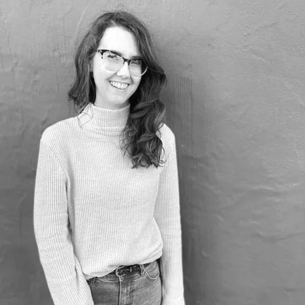 Young woman with long wavy hair and glasses smiling, wearing a turtleneck sweater and jeans, standing in front of a plain wall.