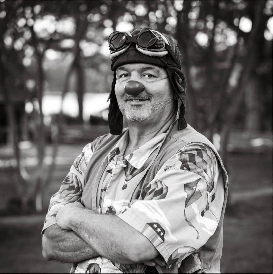 A middle-aged man dressed as a clown, wearing a Hawaiian shirt, with a nose and goggles on his forehead, standing outdoors with trees in the background.