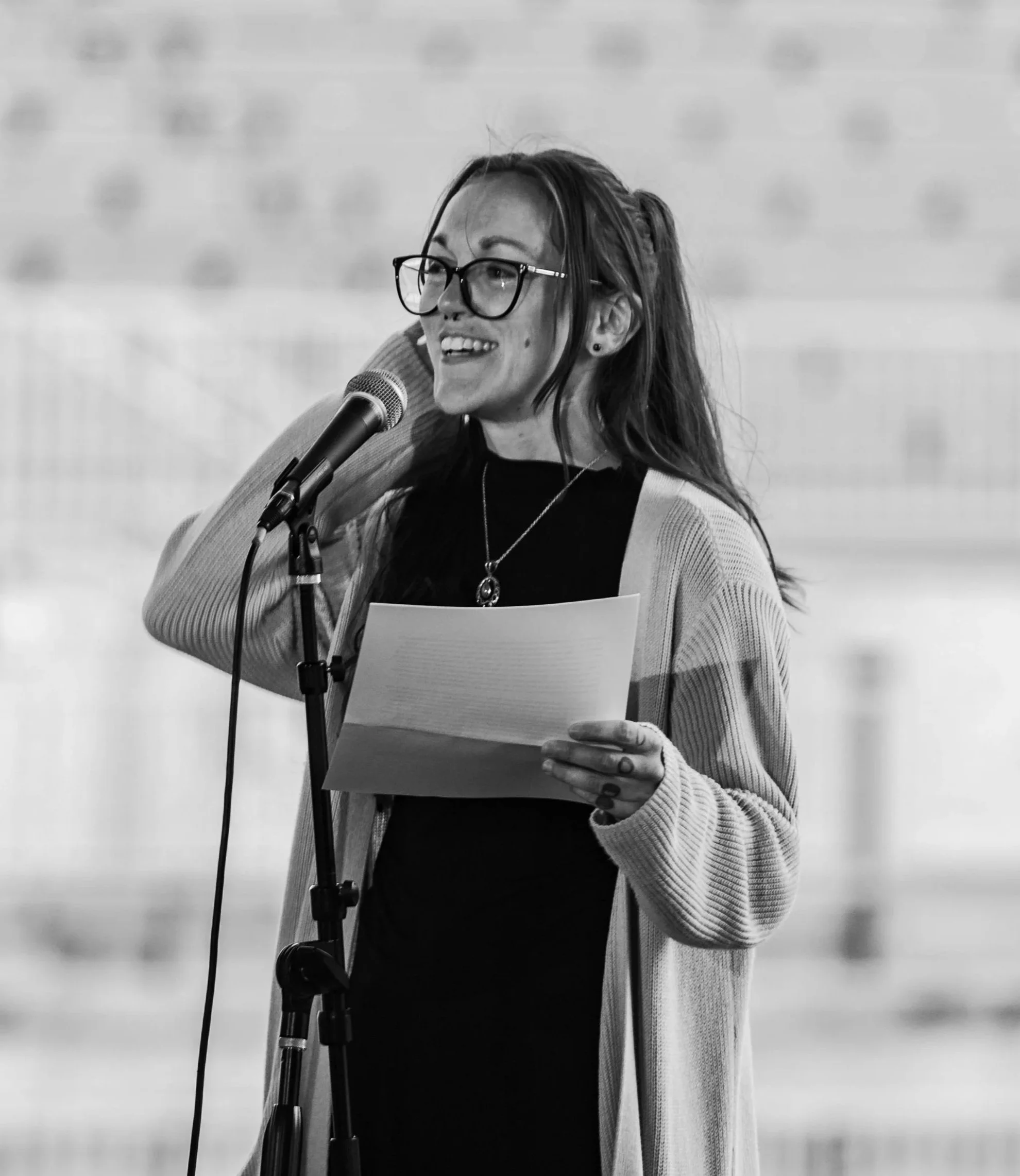 A woman with glasses, long hair, and a necklace, holding a piece of paper, speaking into a microphone, with a happy expression.